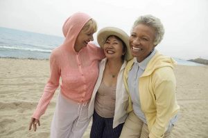 Three smiling older women walking on the beach with their arms around each others' shoulders